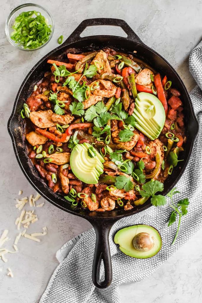 Cast iron photo of a chili recipe with avocado and cilantro toppings.