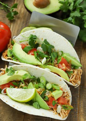Two taco shells filled with slow cooker taco chicken and topped with avocado, tomato and green onions.