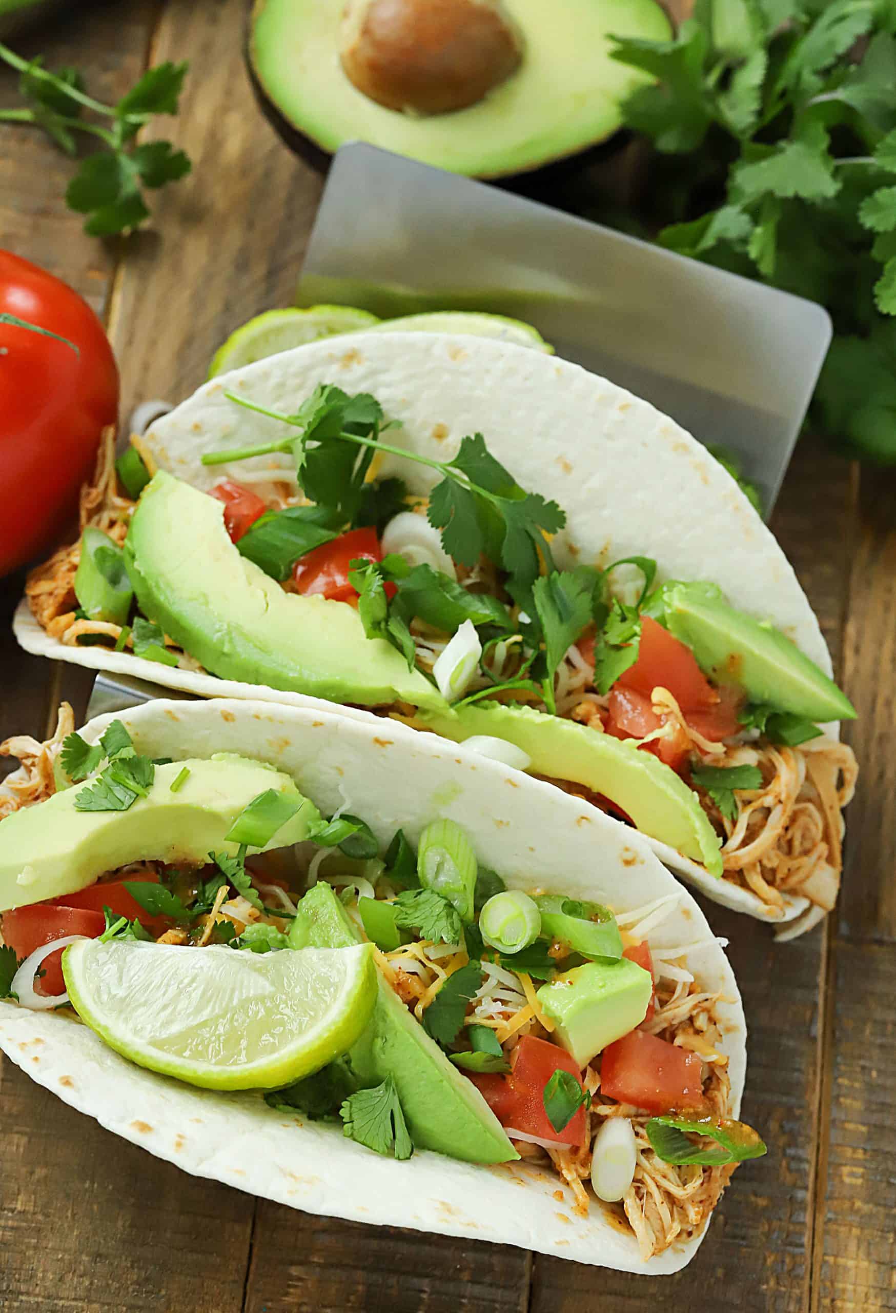 Two taco shells filled with slow cooker taco chicken and topped with avocado, tomato and green onions.